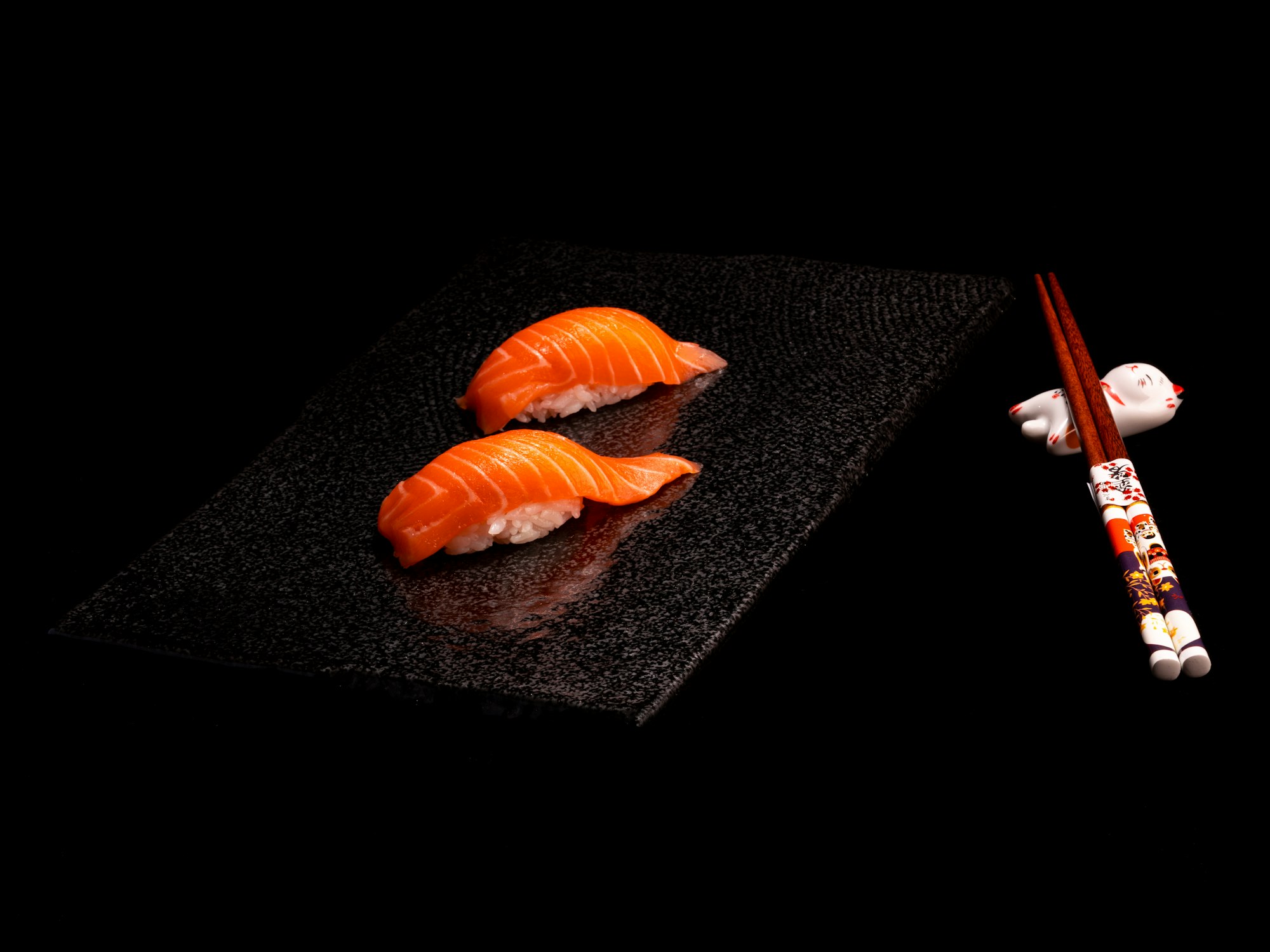 Two pieces of salmon nigiri on a dark slate beside chopsticks